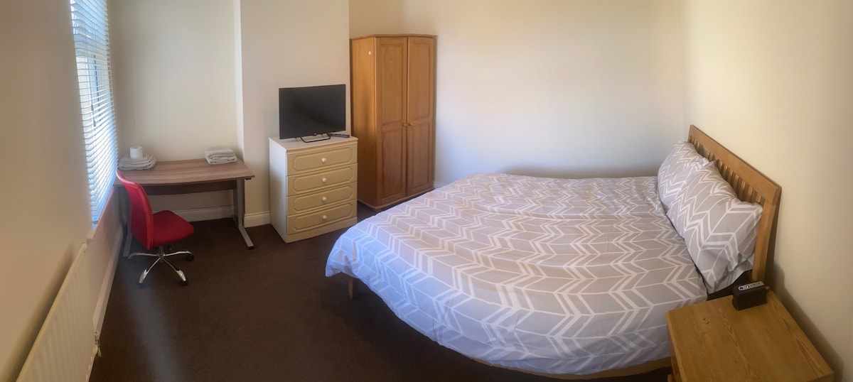 A comfortable double bedroom features a spacious bed with a patterned duvet cover. A wooden wardrobe and a dresser provide ample storage. A desk with a red chair and a television add functionality to the room. Natural light filters through the window with blinds.