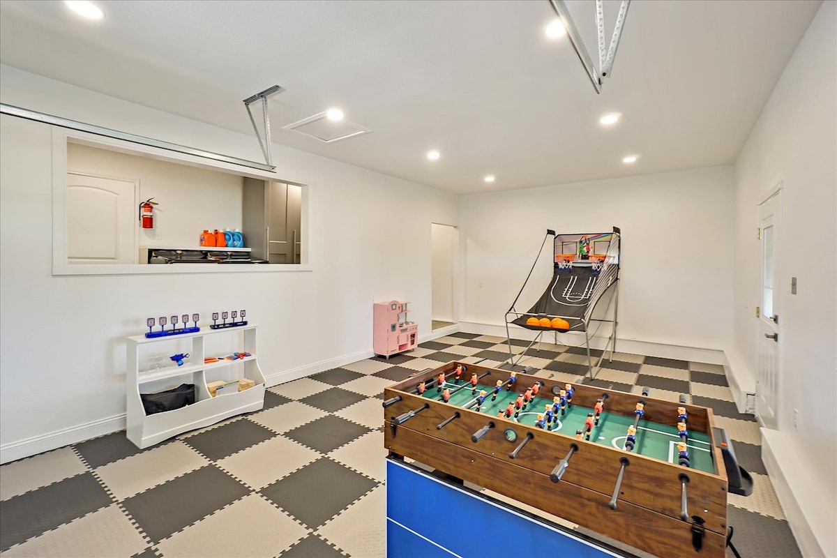 An indoor entertainment area is designed for recreational activities. A foosball table stands prominently, accompanied by a basketball shooting game in the background. Bright lighting illuminates the space, which features a checkerboard floor pattern. A small kitchenette area is visible through an opening.