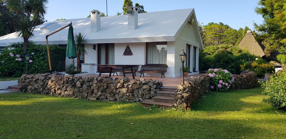 A charming single-story home is surrounded by greenery and vibrant flowers. A spacious wooden deck with seating is visible in front, accompanied by a stone pathway leading to the entrance. The exterior features white brick walls and a sloped roof, enhancing the serene garden setting.