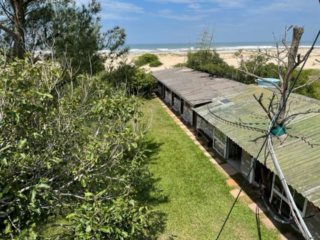 An aerial view of a quaint accommodation surrounded by lush greenery and a manicured lawn. The structure has a rustic roof and is positioned close to the beach, with sandy shores visible in the background under a clear blue sky.
