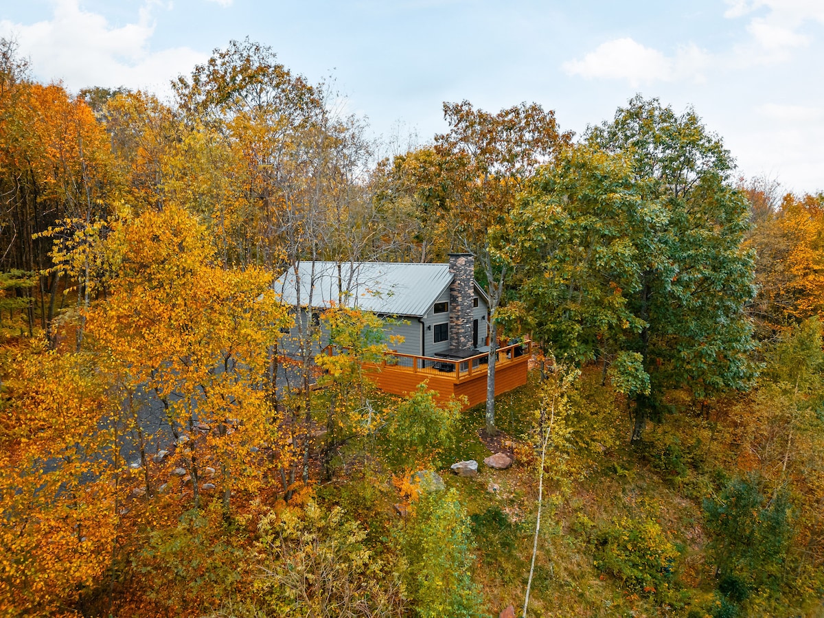 A cabin nestled among trees adorned with autumn foliage. The exterior features a deck and a chimney, blending harmoniously into the surrounding nature. Sunlight filters through the leaves, creating a serene setting ideal for relaxation.