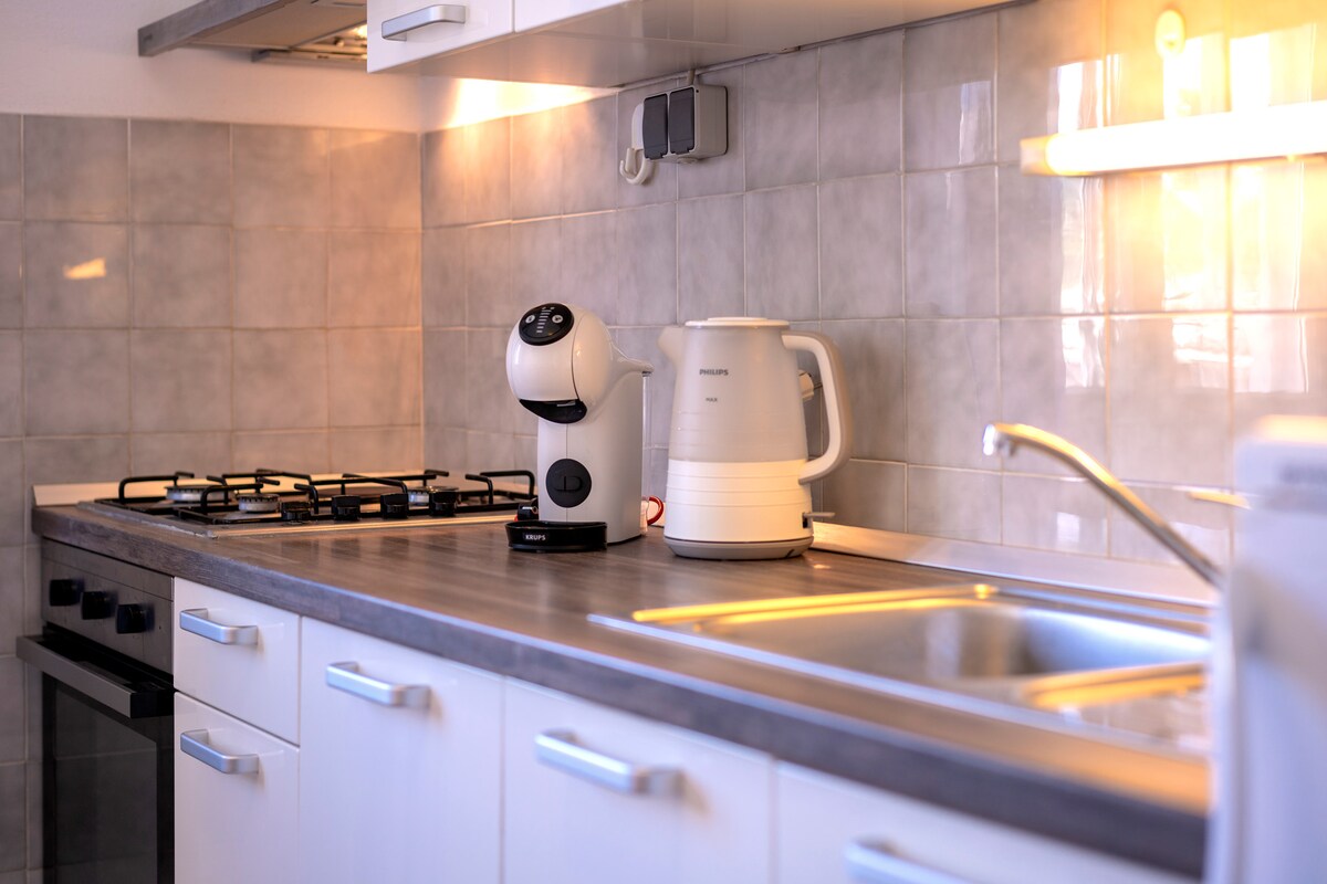 A modern kitchen area is shown, featuring a sleek countertop with a gas stove and an oven. A coffee maker and an electric kettle are positioned on the counter. Light-colored tiles adorn the walls, enhancing the clean and functional layout.