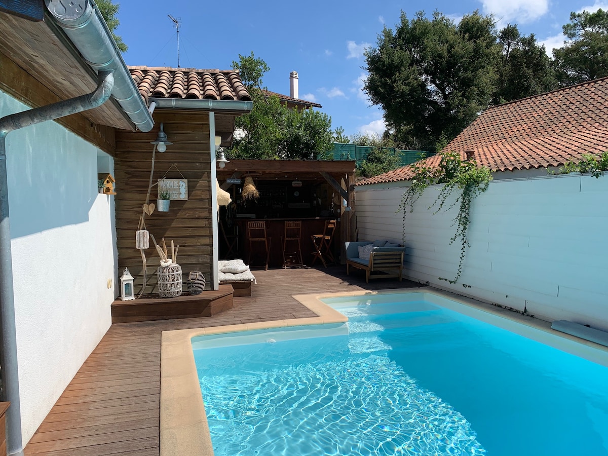 A wooden deck surrounds a clear swimming pool, reflecting the blue sky above. An outdoor bar area with seating is positioned under a shaded structure. Lounge chairs are arranged nearby, creating a relaxed atmosphere, while greenery adds a natural touch to the surroundings.