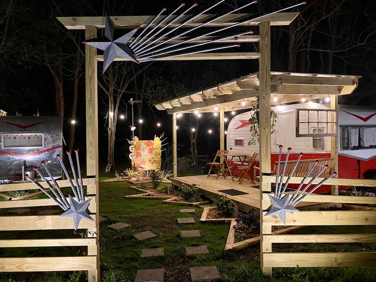 A charming entrance to the Glam Camp is illuminated at night, featuring decorative stars and lights. The pathway is lined with stepping stones leading to vintage campers, surrounded by green grass and trees, creating a welcoming atmosphere for guests.