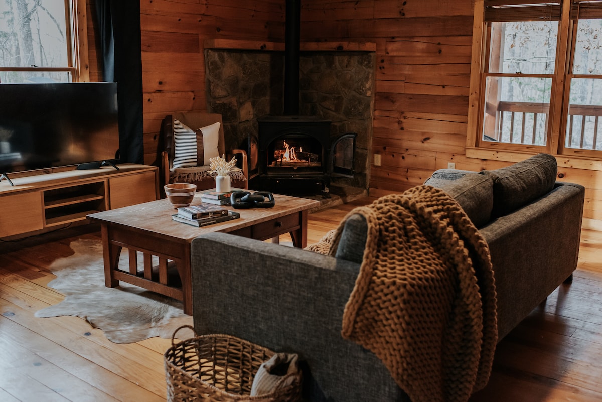A cozy living room features wooden walls and a stone fireplace, providing warmth and comfort. A gray sectional sofa is complemented by a textured throw and a wooden coffee table with books. Large windows allow natural light, while a TV is positioned nearby for entertainment.