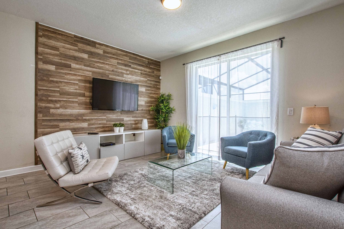 A modern living area is highlighted by a textured wood accent wall and a flat-screen television. Comfortable blue chairs and a gray sofa provide seating options, while a glass coffee table sits on a soft area rug. Natural light filters through sheer curtains, enhancing the inviting space.