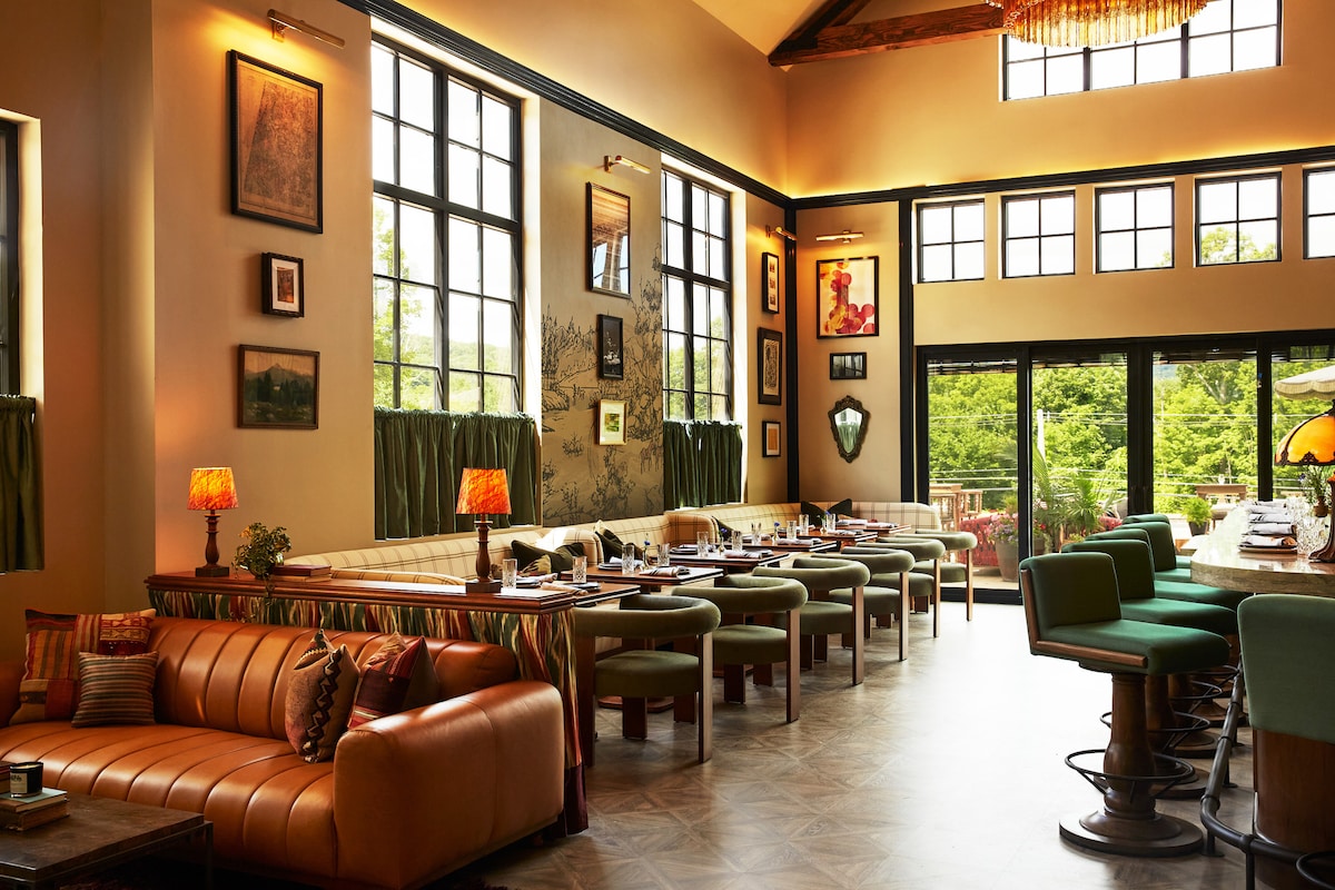 A spacious dining area is presented with large windows allowing natural light to illuminate the space. Comfortable seating is arranged around wooden tables, accentuated by various artworks and decorative lamps. Green curtains complement the earthy tones of the furnishings.