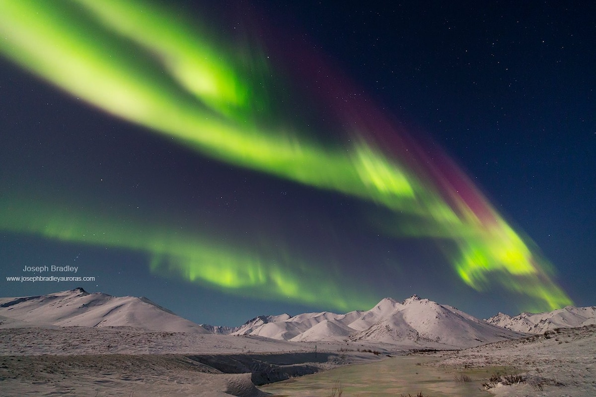 The night sky is filled with vibrant green and purple hues of the Northern Lights, illuminating the mountain landscape below. Snow-covered hills and a dark foreground create a serene contrast against the colorful aurora borealis, enhancing the natural beauty of the wilderness.