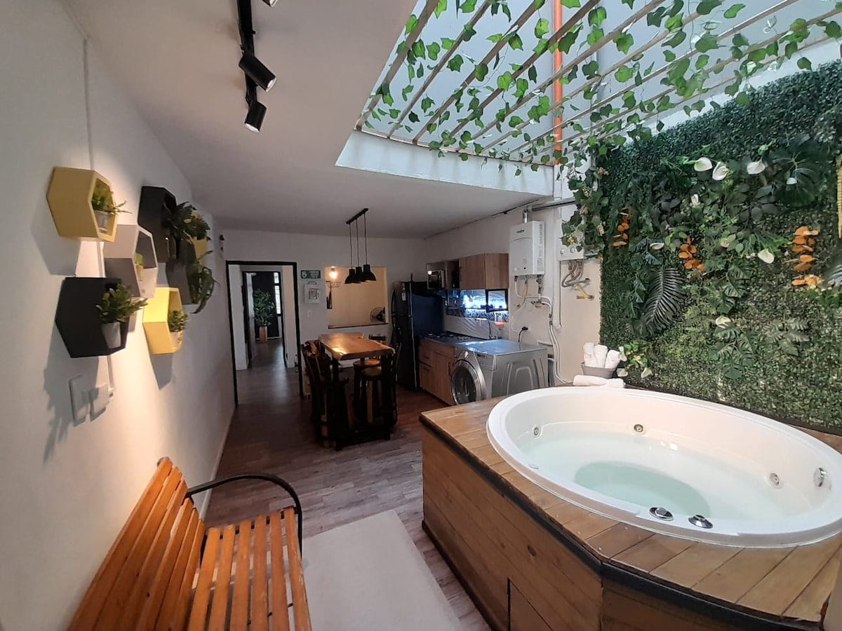 A modern living space is appointed with a hot tub, surrounded by greenery and decorative wall planters. A wooden dining table is visible, accompanied by a well-equipped kitchen in the background. Natural light enters through a skylight, enhancing the room's openness.