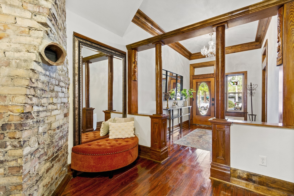 An inviting entryway showcases elegant woodwork and a mixture of textures. A curved, upholstered bench in warm tones provides seating, while large mirrors enhance the sense of space. Natural light filters through the front windows, highlighting the charm of the historic architecture.
