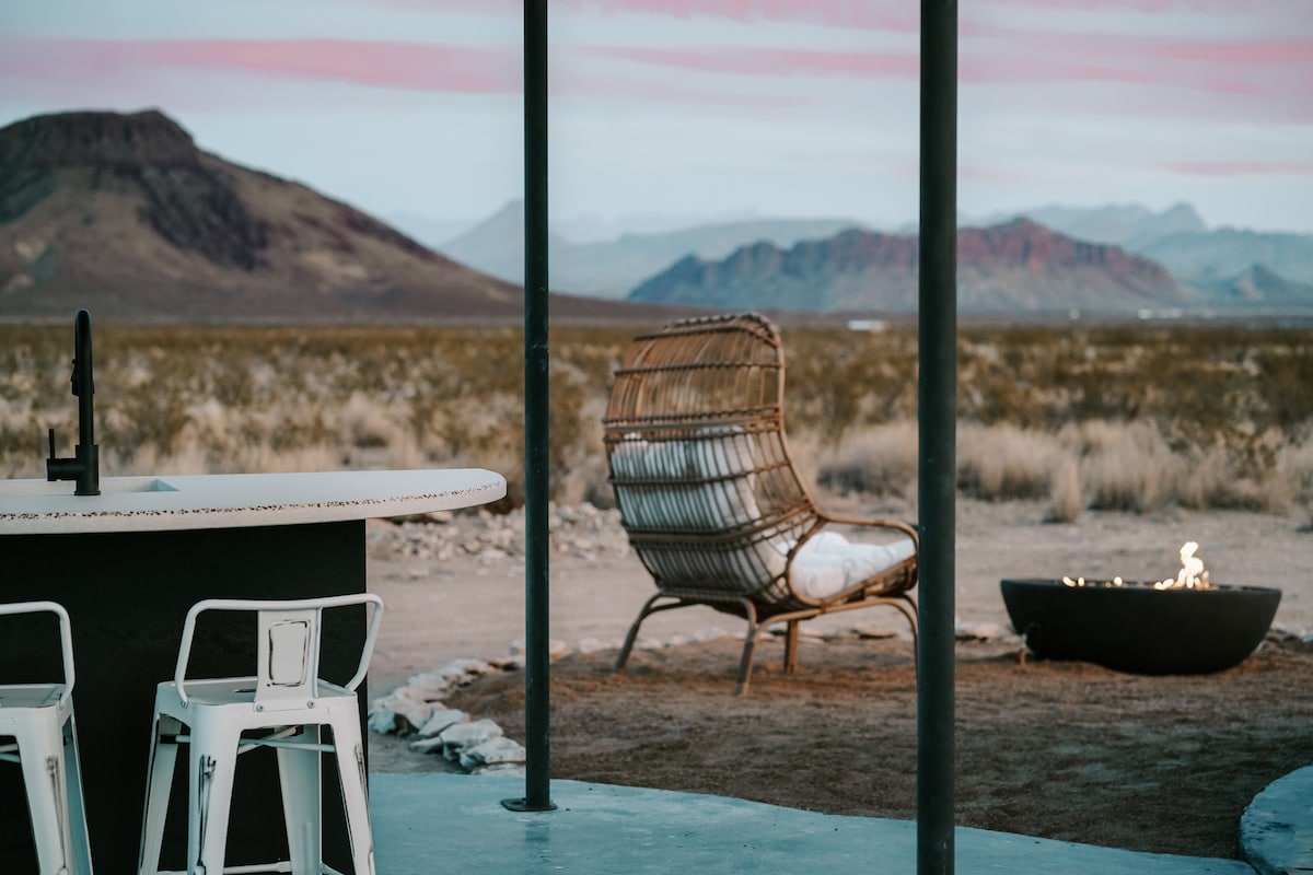 An outdoor seating area is visible, featuring a woven lounge chair positioned near a fire pit. A circular dining table with metal chairs is situated nearby, surrounded by expansive desert views and distant mountains under soft evening hues.