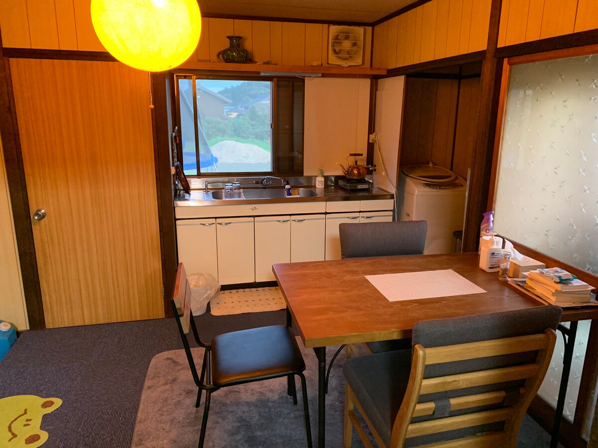 A modest kitchen area features a wooden dining table with four chairs positioned on a blue carpet. A large window provides natural light, while a sink and stove are visible in the kitchen space. Laundry facilities are located in the corner.