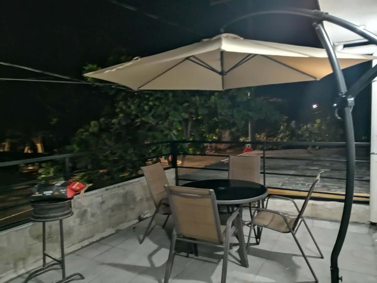 An outdoor terrace is presented with a round table surrounded by four chairs. A large umbrella provides shade, and a tall bar table is visible. The setting is illuminated softly, revealing greenery in the background amidst the serene evening atmosphere.
