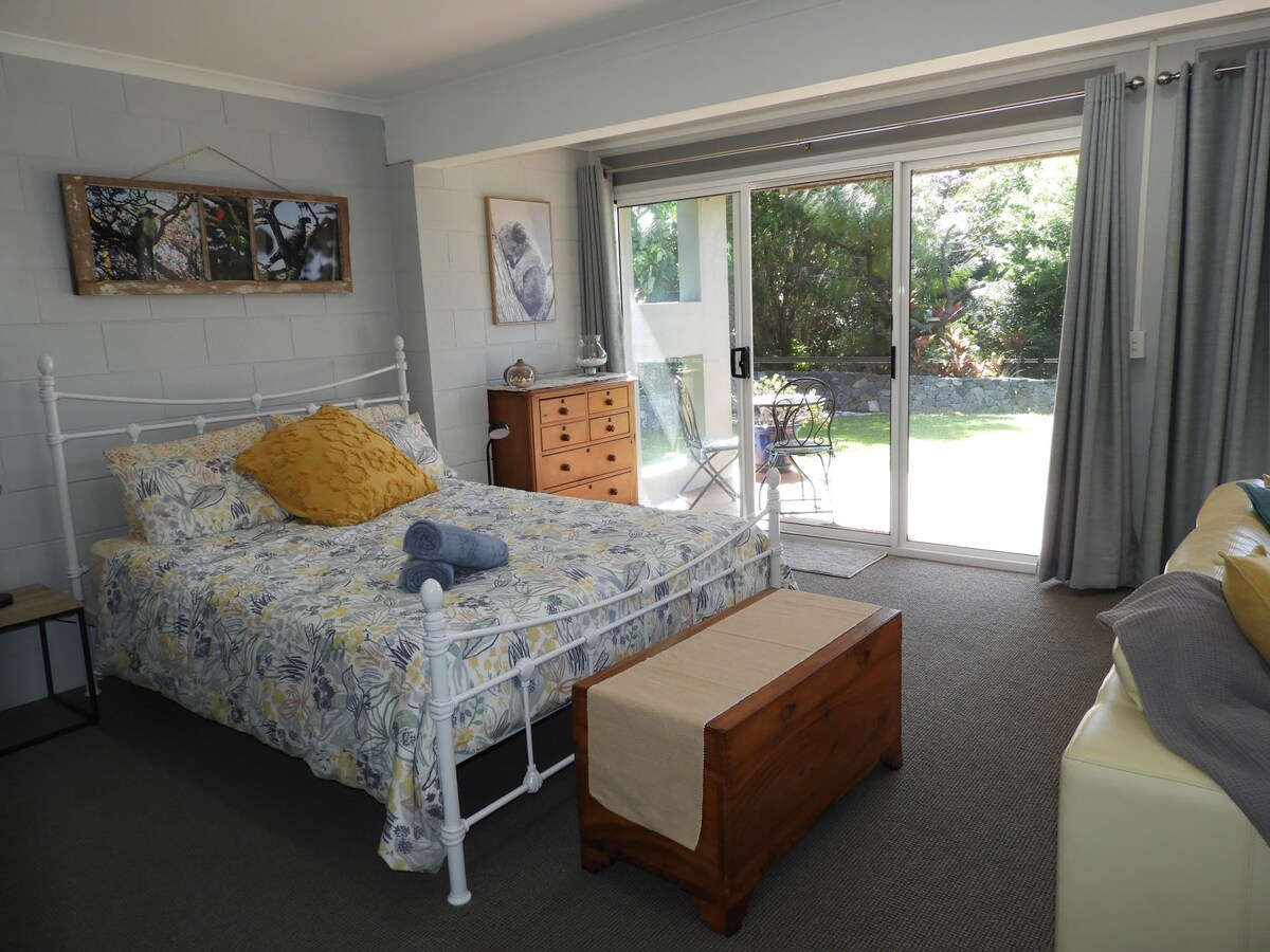 A comfortably arranged sleeping area features a white metal bed with a floral-patterned quilt and a coordinating yellow pillow. A wooden chest sits at the foot of the bed, while a sliding glass door opens to a private patio and garden view.