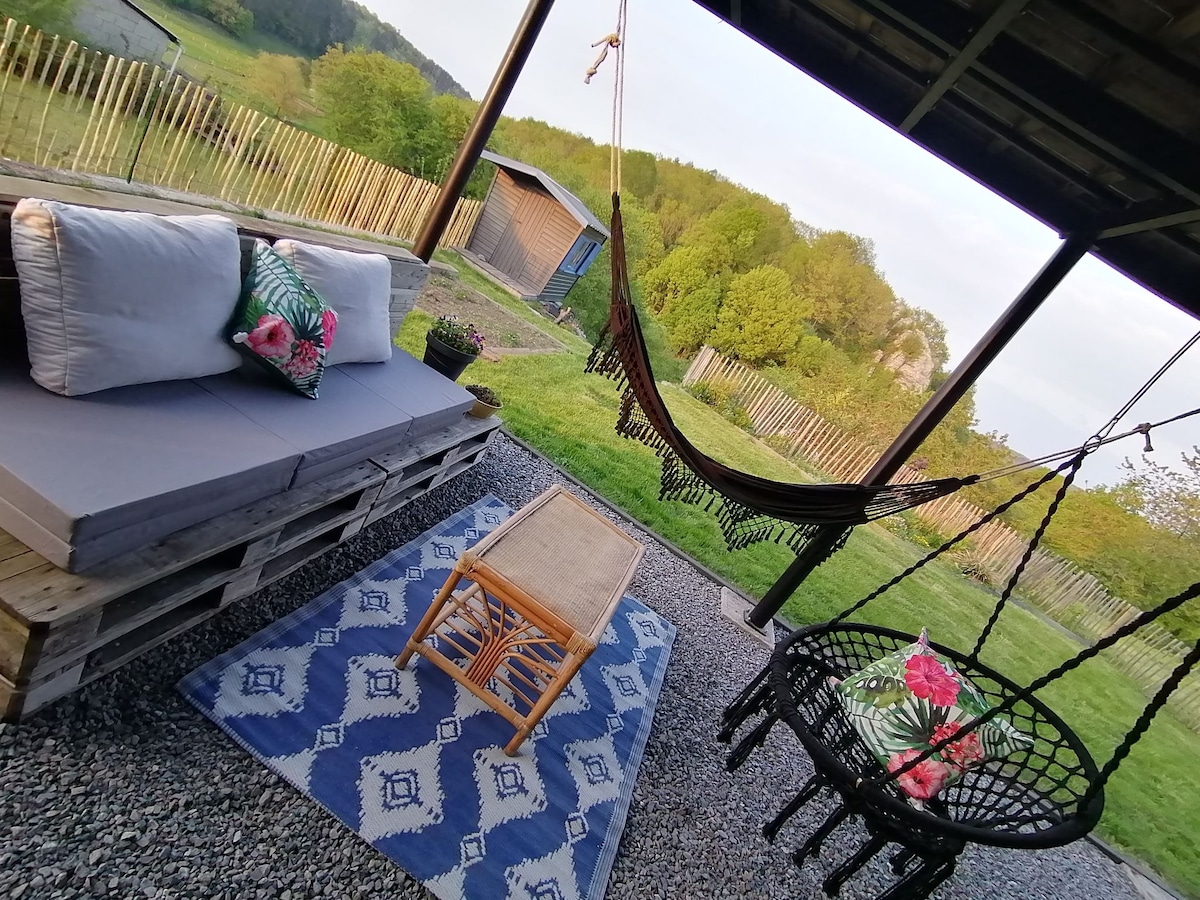 An outdoor seating area is arranged with a hammock, a hanging chair, and a low coffee table atop a decorative outdoor rug. The setting overlooks a green landscape, featuring wooden fences and a distant shed surrounded by nature.