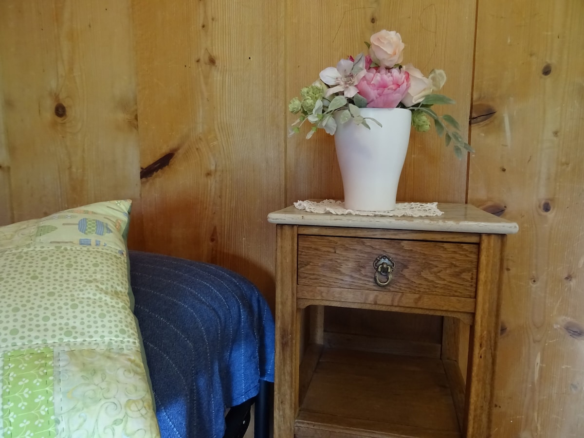 A wooden nightstand with a marble top and a decorative flower vase sits beside a cozy bed. The wall is made of warm wood panels, complementing the green and blue patterned bedding. A delicate lace doily rests on the nightstand.
