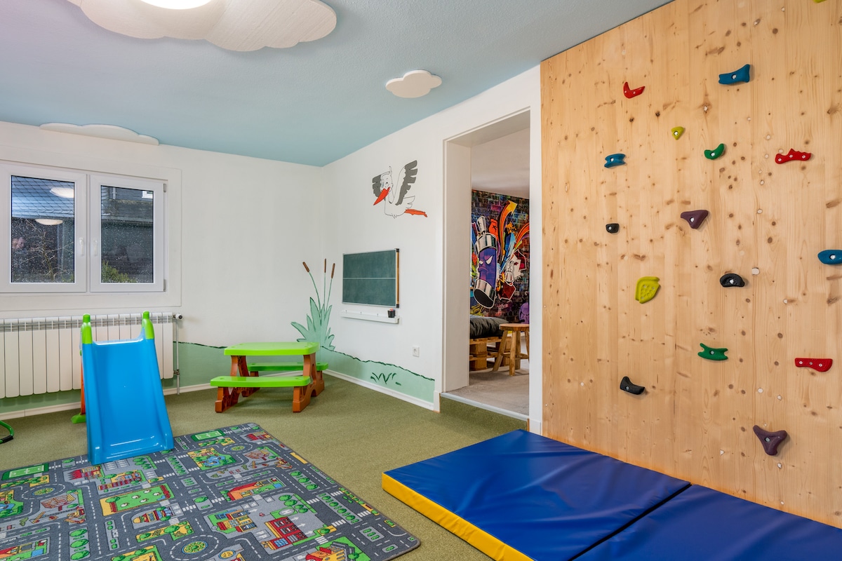 A vibrant indoor play area features a small slide and a climbing wall. Colorful floor mats are laid out, depicting roads and buildings. The walls display playful murals, contributing to a lively and engaging environment suitable for children.