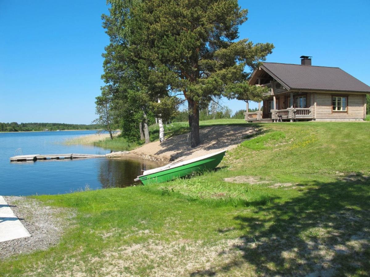 A wooden cabin is situated near the water's edge, surrounded by greenery. A small green boat rests on the shore, while the calm lake reflects the clear blue sky. A spacious deck with outdoor seating is visible at the cabin's entrance.