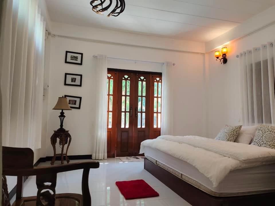 A serene bedroom features a spacious bed with crisp white bedding, positioned near large wooden doors that let in natural light. Soft curtains frame the windows, and a small red rug adds a touch of color. A bedside lamp and decorative frames enhance the atmosphere.