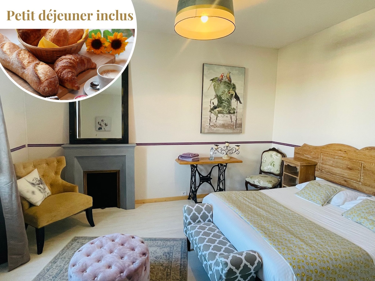 A spacious room features a comfortable bed with light bedding and decorative pillows. A plush ottoman sits at the foot of the bed. A small table is positioned beside a chair, with a piece of art displayed on the wall. Natural light enters through a window.