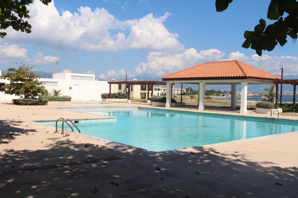 A spacious outdoor pool is framed by a sunlit deck and shaded areas. A covered pavilion with a terracotta roof sits adjacent to the pool. Lush landscaping and distant views of the ocean are visible under a clear blue sky with scattered clouds.