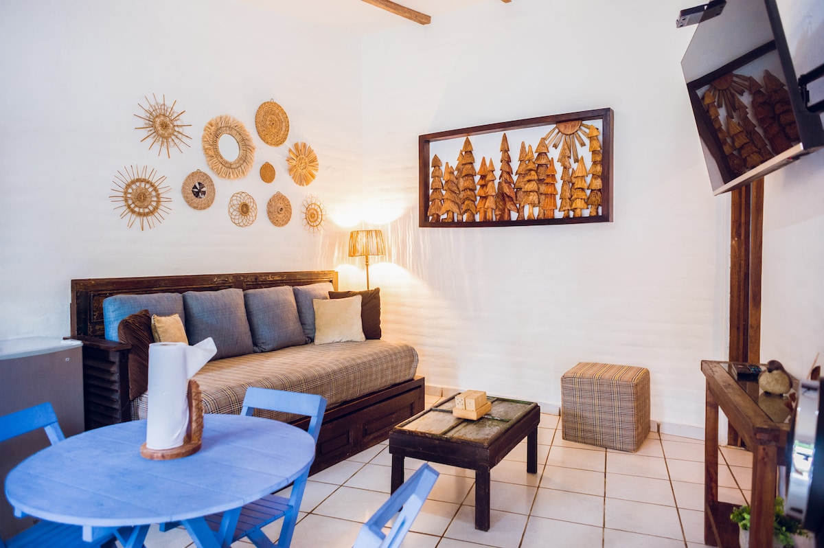An inviting seating area features a daybed adorned with decorative pillows, complemented by a textured wooden coffee table. A light fixture provides warmth, while a collection of handcrafted art pieces hangs on the wall. A blue dining table with chairs creates a vibrant corner.