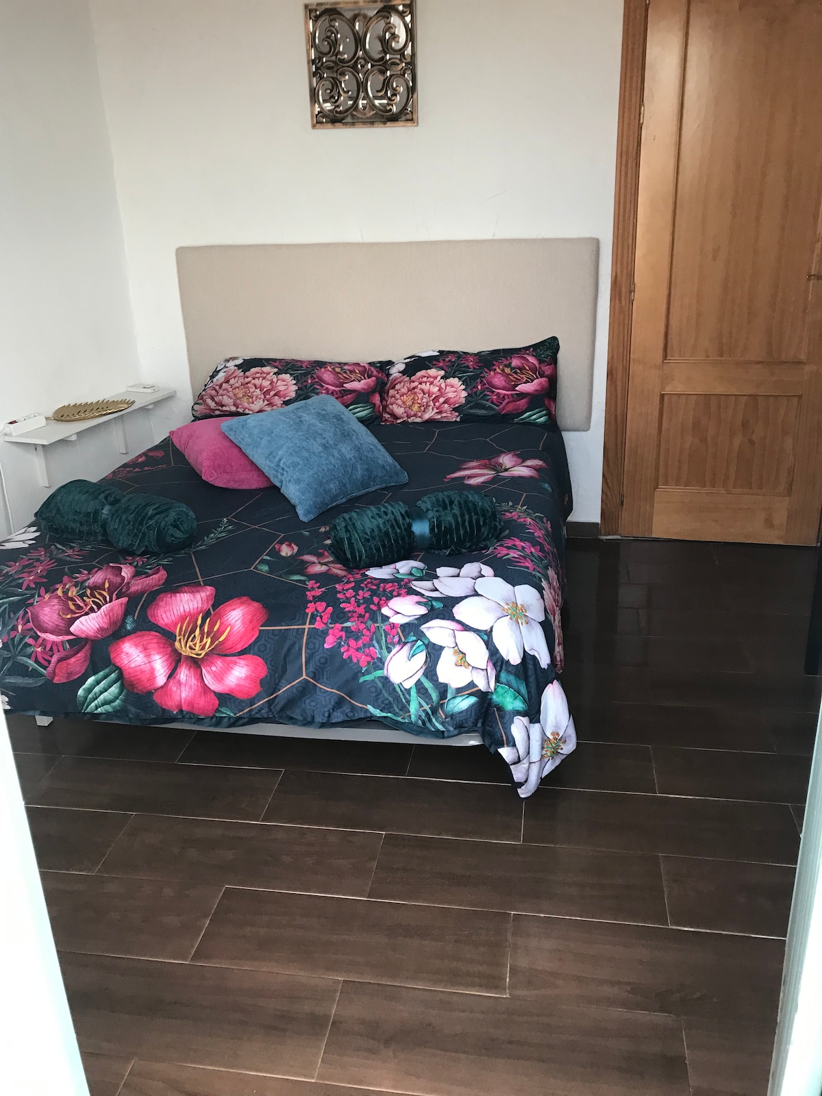 A cozy bedroom is furnished with a bed featuring a floral quilt and accent pillows in teal and blue. Wooden flooring complements the soft hues of the wall. A small nightstand is located beside the bed, and a wooden door leads outside.