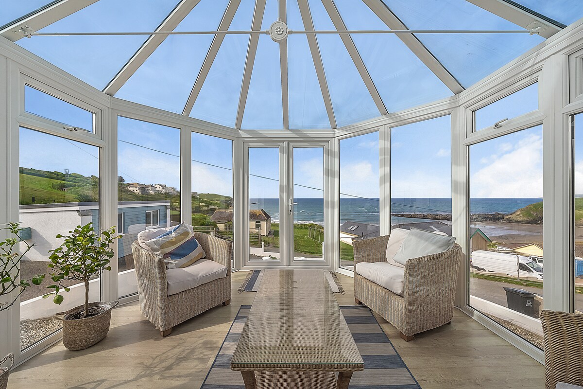 A bright conservatory is shown with large glass windows offering panoramic views of the coastline and sea. Two wicker armchairs are positioned around a wooden coffee table, creating a cozy space to relax and enjoy the scenery. Natural light fills the area, enhancing the inviting atmosphere.