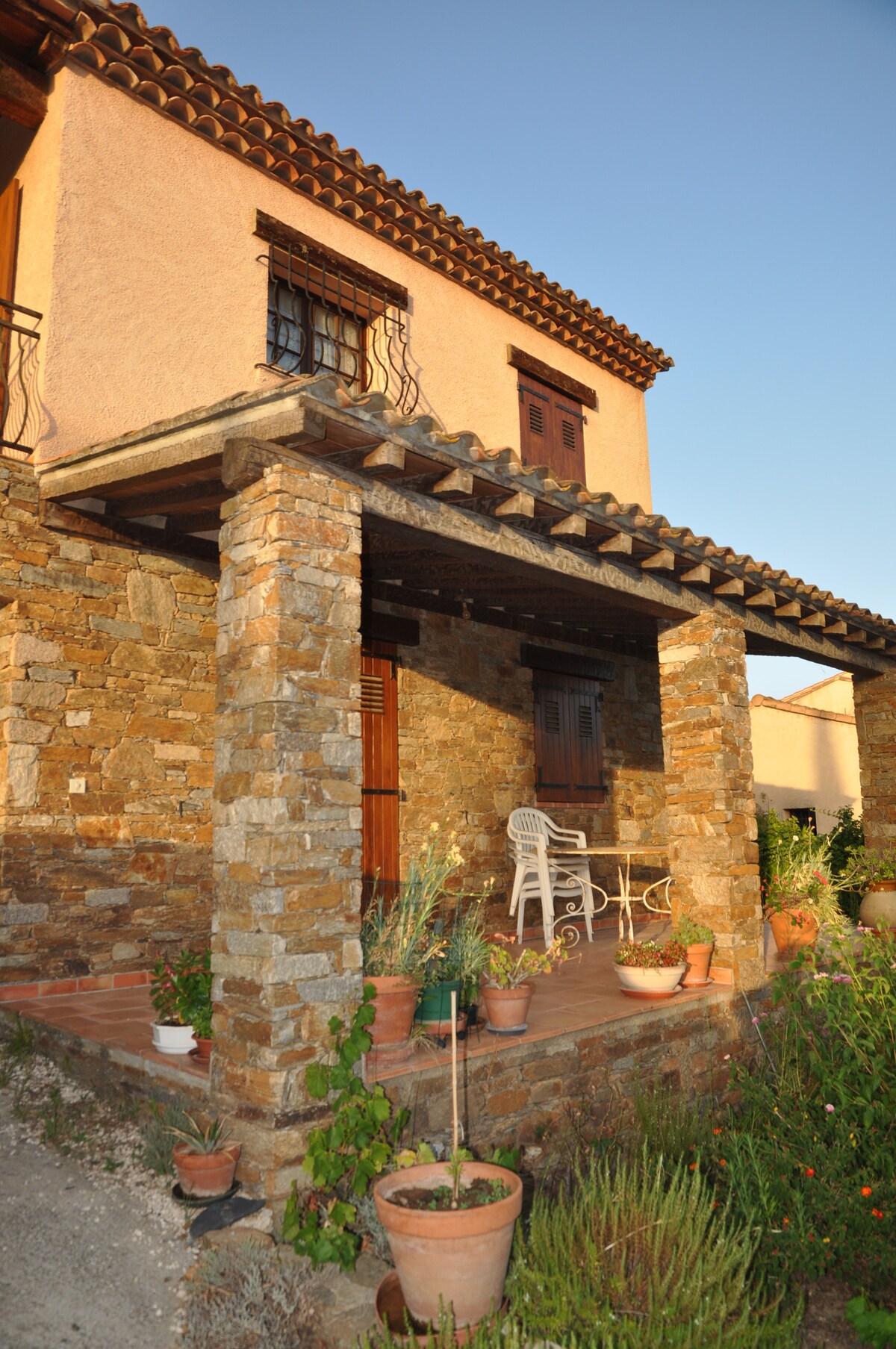 The exterior features a charming stone facade with a covered porch. Potted plants line the entrance, and a white chair and small table are positioned under the overhang. A wooden door leads into the house, complemented by wooden window shutters above.