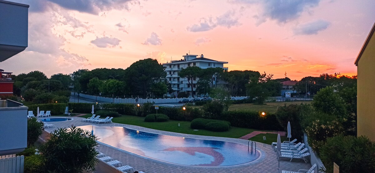 An outdoor pool is surrounded by neatly arranged lounge chairs and landscaped greenery, with the sunset casting warm hues across the scene. Buildings can be seen in the background, framed by trees and a peaceful sky, creating a serene environment.
