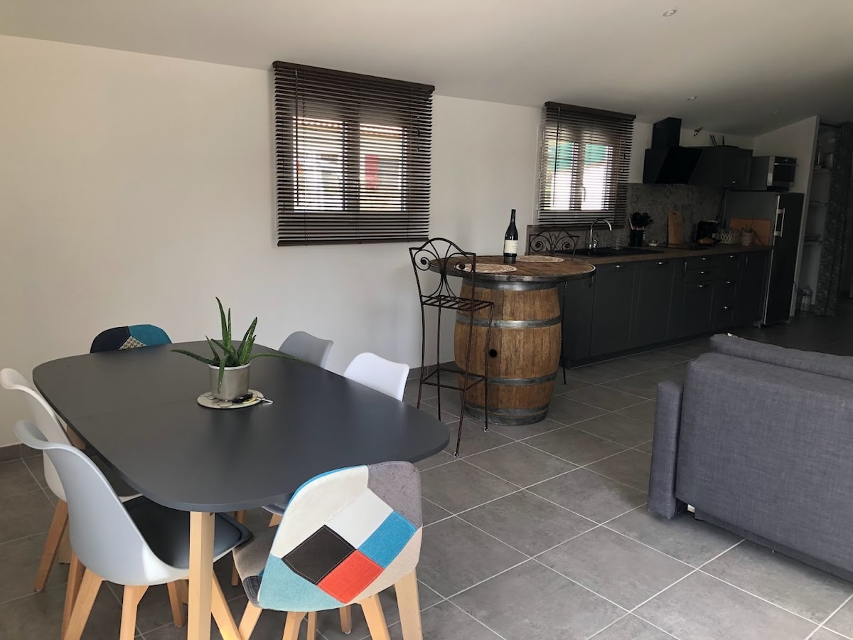 A spacious kitchen and dining area features a contemporary table surrounded by multi-colored chairs. A large barrel serves as a unique focal point, while modern appliances and cabinetry create a functional atmosphere. Natural light enters through the windows, enhancing the open layout.