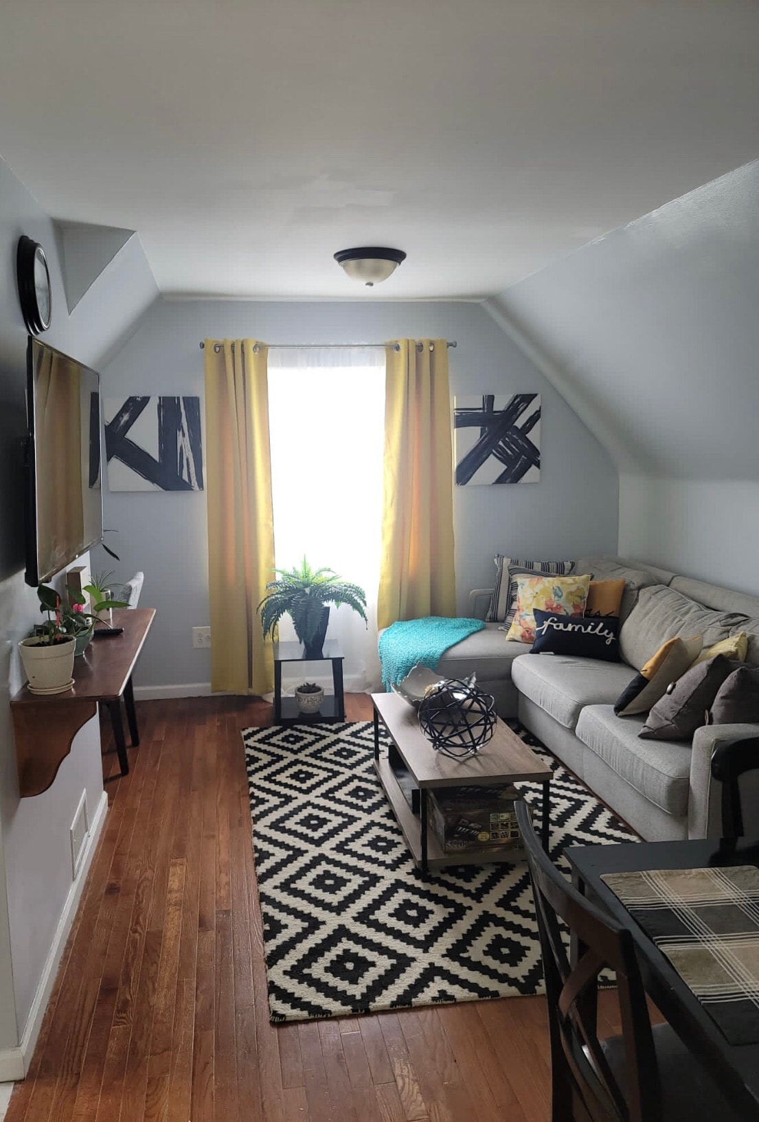 A cozy living area features a sofa adorned with decorative cushions, accompanied by a coffee table. A vibrant area rug sets the stage for a warm atmosphere. Natural light streams through the window, framed by yellow curtains, while potted plants add a touch of greenery.