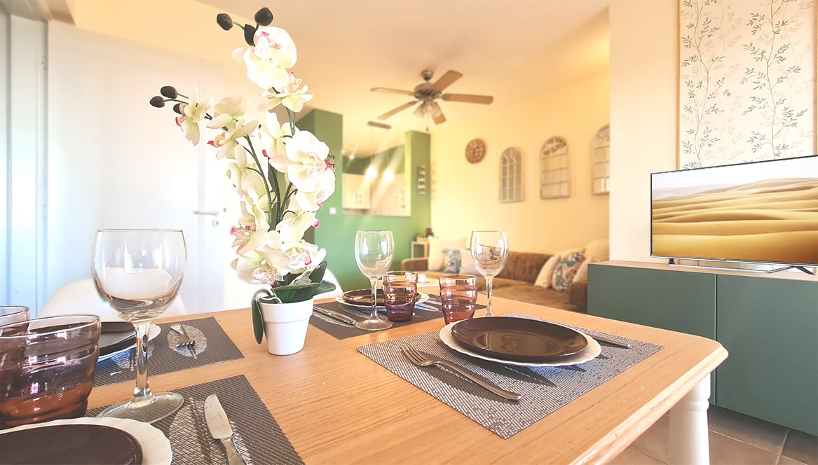 A dining table is set with plates, cutlery, and glassware, complemented by a decorative plant. In the background, a comfortable lounge area is visible, featuring a smart TV and light-colored furniture. Natural light fills the space, enhancing the open plan layout.