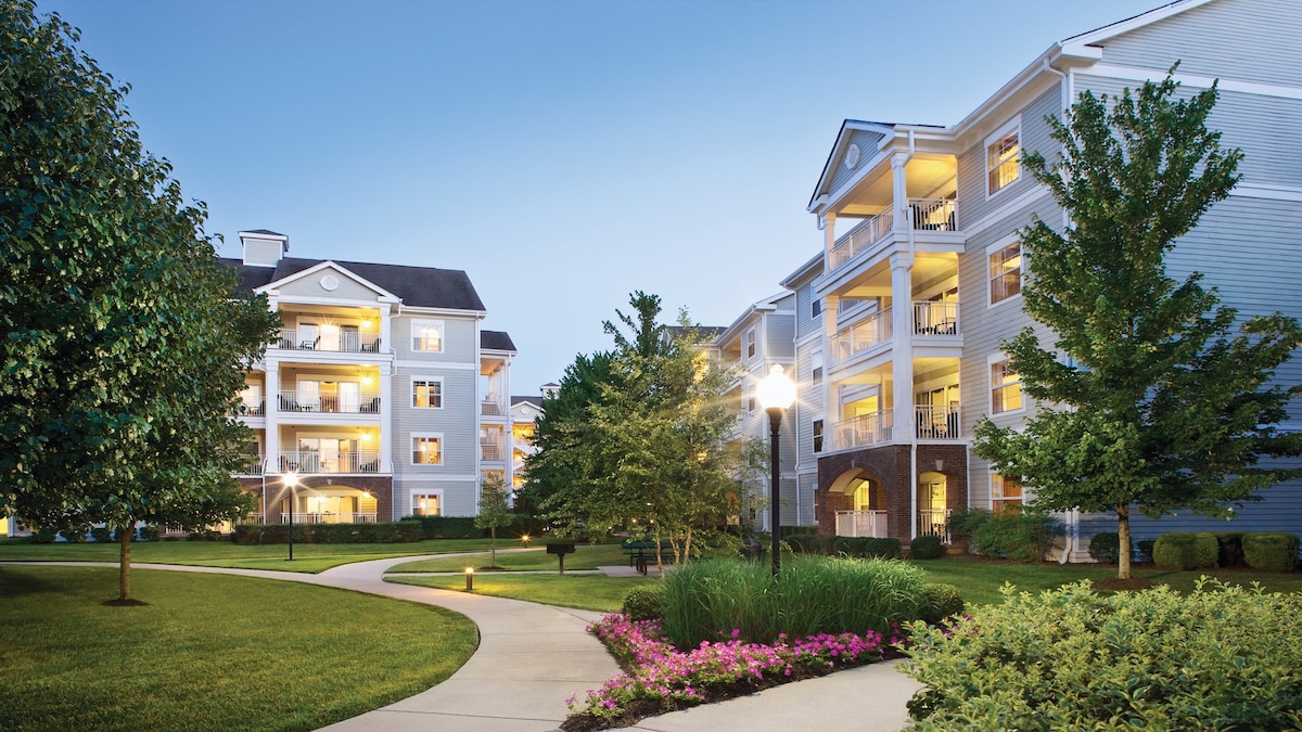 A well-maintained green space surrounds the buildings, featuring neatly trimmed grass and blooming plants. Pathways meander through the landscape, leading to the entrances of the multi-story condos. Soft lighting from lamps highlights the welcoming atmosphere in the evening.