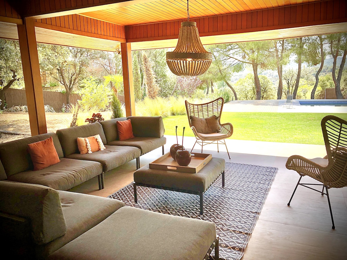 An indoor seating area features a comfortable arrangement of plush sofas and armchairs. A central coffee table holds decorative items. Large sliding doors provide a view of the garden and pool, allowing natural light to fill the space.