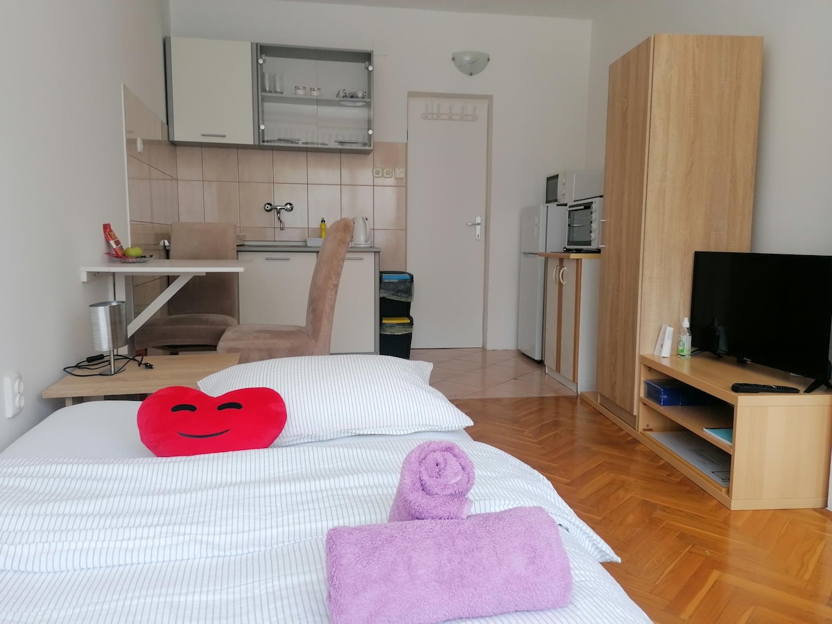 A comfortable sleeping area features neatly arranged bedding, accented by a bright red heart-shaped pillow. A small kitchenette is visible in the background, accompanied by a dining space with a light-colored chair. A television sits atop a wooden stand beside a storage cabinet.