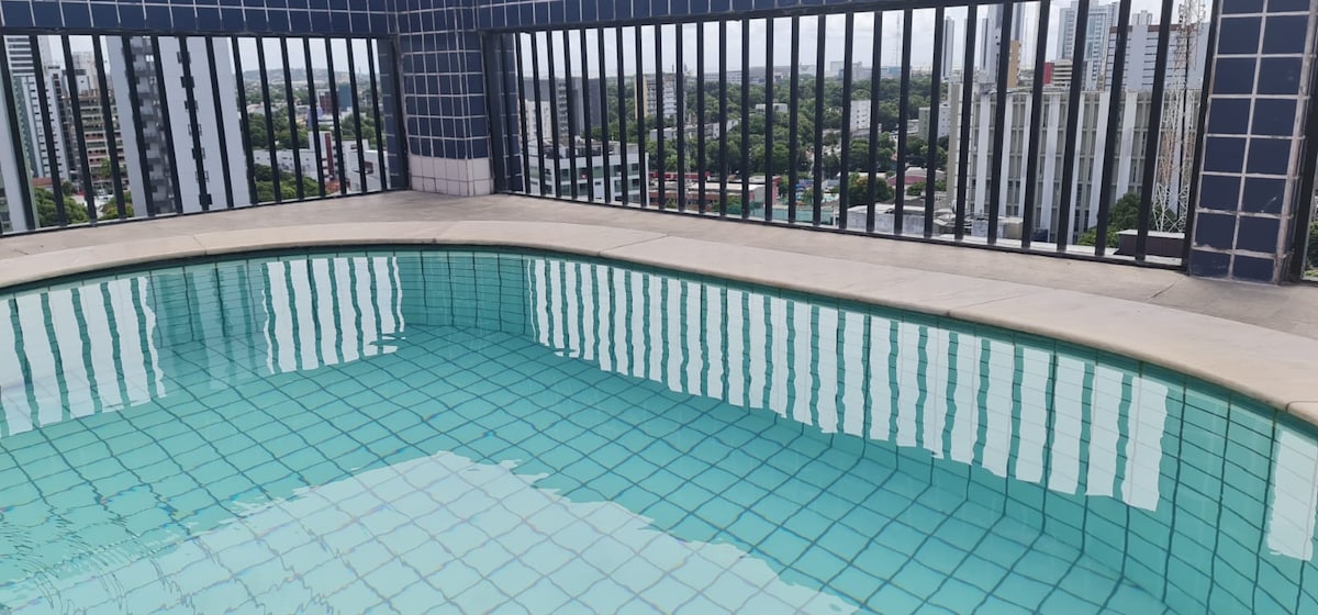 A rooftop pool is viewed, featuring clear water reflecting the surrounding city skyline. The pool's edges are outlined with light-colored tiles, and the space is surrounded by black railings, offering an unobstructed view of the greenery and buildings in the distance.