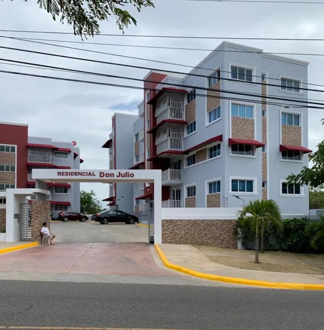 A modern residential complex is presented, featuring three stories with a mix of brick and stucco finishes. A gated entrance is visible, alongside landscaped areas with greenery. Several windows reflect natural light, and a paved driveway leads into the property.