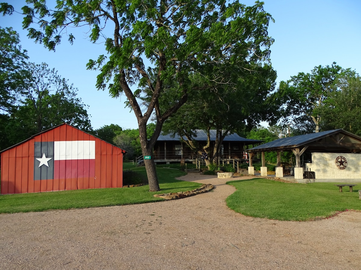 A scenic outdoor view captures a vibrant red barn adorned with a Texas flag alongside two inviting houses and a pavilion. Lush green lawns are surrounded by trees, creating a serene atmosphere. A gravel path winds through the property, leading to various gathering spaces.