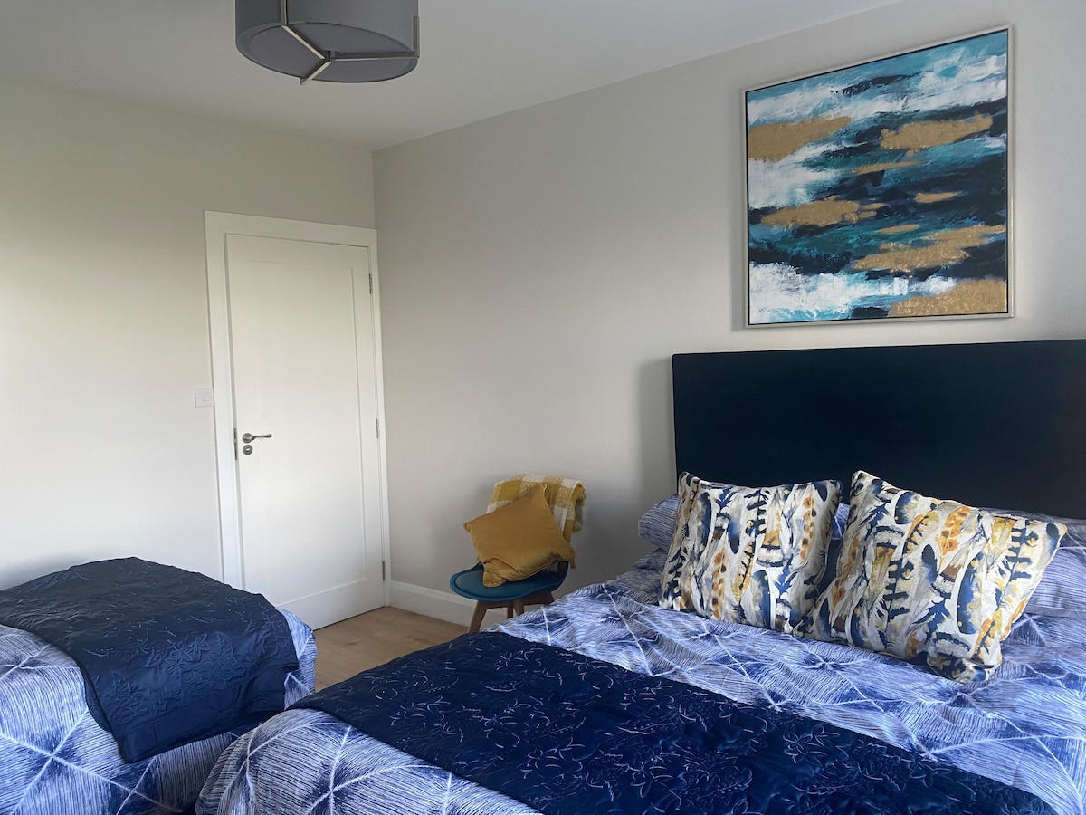 A spacious bedroom is furnished with a king size and single bed, both dressed in patterned bedding. A decorative painting adorns the wall above the beds. A door leads to the exterior, and a small table with a yellow throw is positioned beside the single bed.