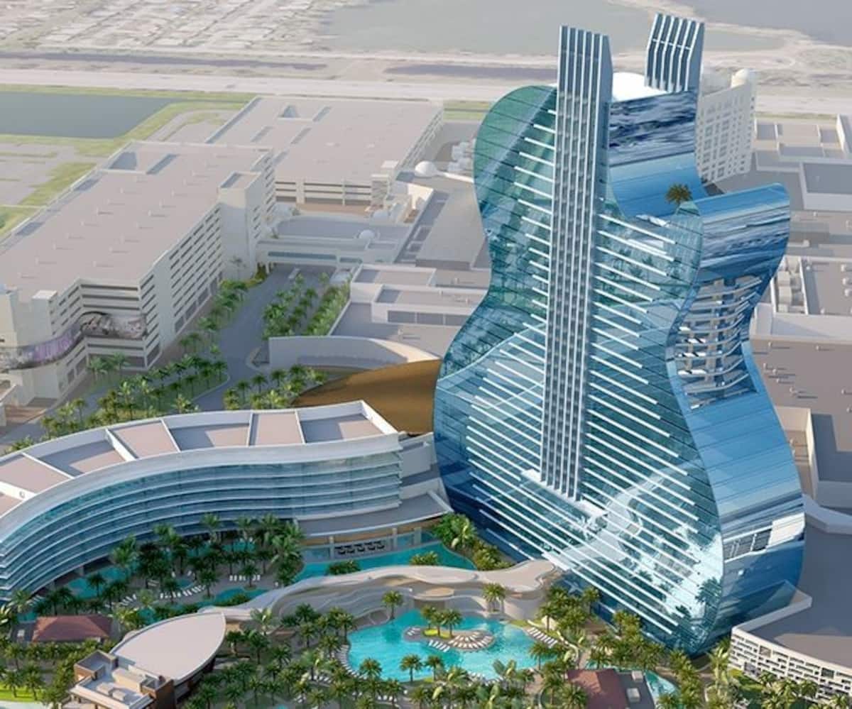The image displays a modern hotel structure designed in the shape of a guitar, with sleek glass facades reflecting the surroundings. Surrounding palm trees and swimming pools contribute to a tropical ambiance. The layout showcases a combination of entertainment and relaxation spaces.