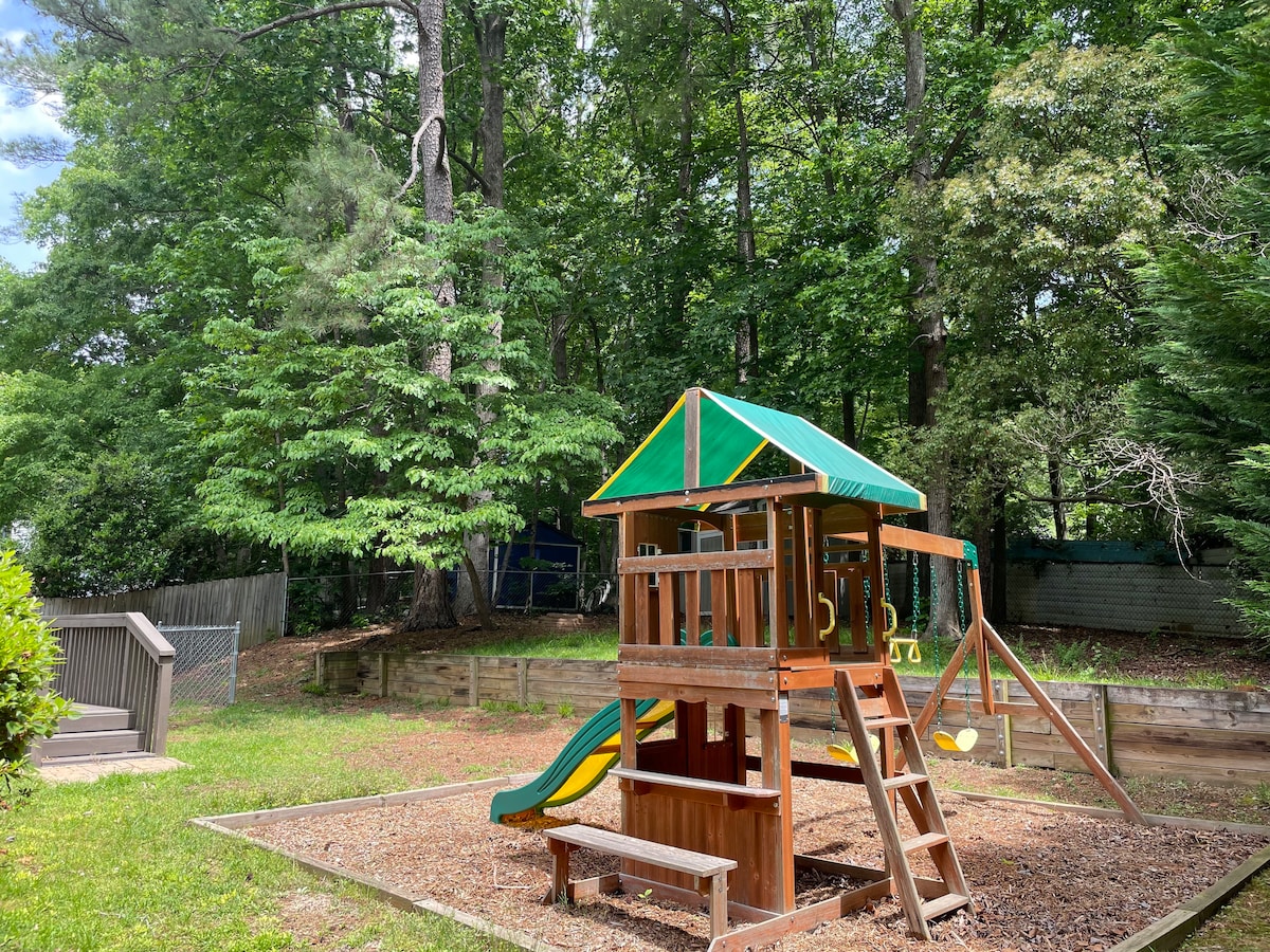A wooden playground structure is positioned within a shaded outdoor area surrounded by tall trees. Features include a slide, swings, and a small climbing area, providing a space for outdoor play. A bench is located nearby, nestled on the wood-chip surface.