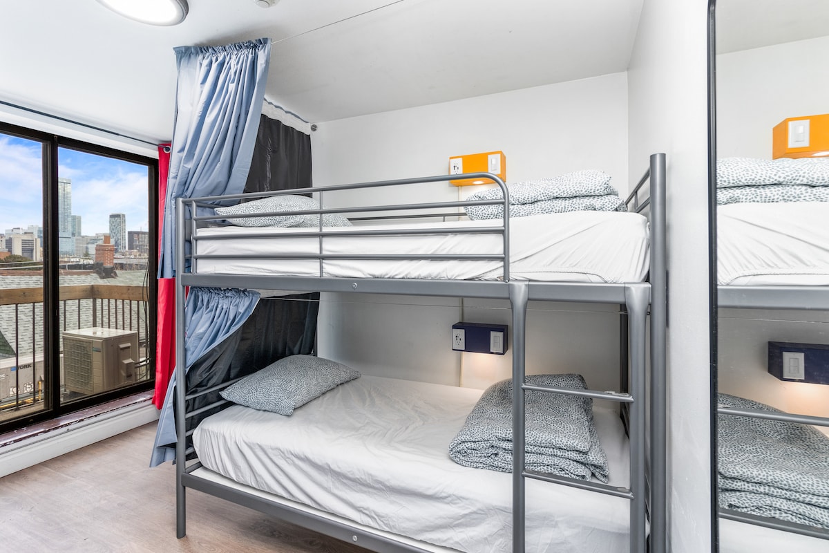 A dorm room features a metal bunk bed with white bedding and navy patterned pillows. A window allows natural light to fill the space, offering views of the surrounding area. Nightstand lamps are positioned beside the lower bunk for convenience.