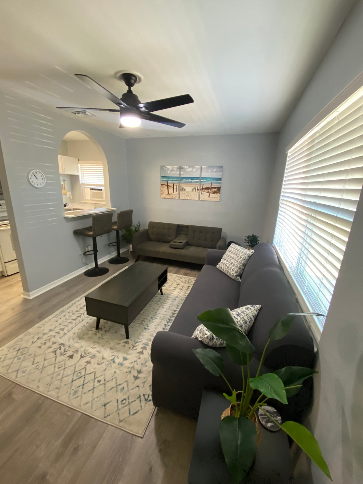 A comfortable living area showcases a dark gray sofa and a matching loveseat, complemented by a coffee table in the center. A decorative rug lies beneath, and a wall clock is positioned nearby. Light streams in through window blinds, illuminating the space.