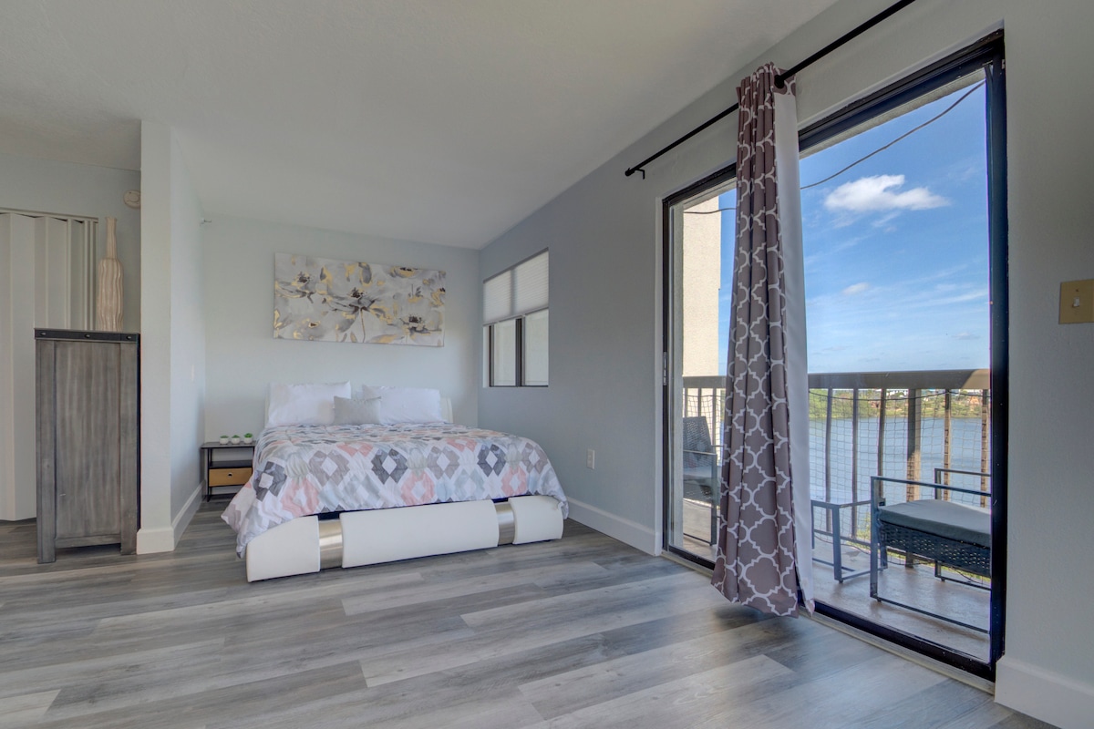 A spacious bedroom is highlighted by a comfortable bed covered with a patterned quilt. Large windows allow natural light to fill the room, while a balcony with seating overlooks a picturesque lake. Modern décor and light-colored walls enhance the tranquil atmosphere.
