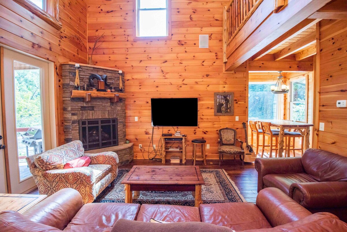 A spacious living area features warm wooden walls and a cozy stone fireplace. Comfortable seating is arranged around a wooden coffee table, with a large television mounted above the fireplace. Natural light floods in through large windows, highlighting the inviting atmosphere.