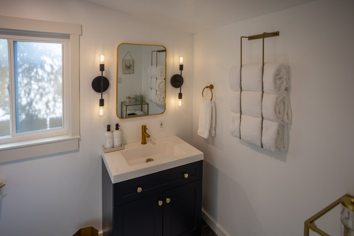 The bathroom features a dark cabinetry unit with a white countertop and gold fixtures. A large mirror is mounted above the sink, complemented by stylish wall sconces. Soft, neatly hung towels are arranged nearby, and natural light filters in through a window.