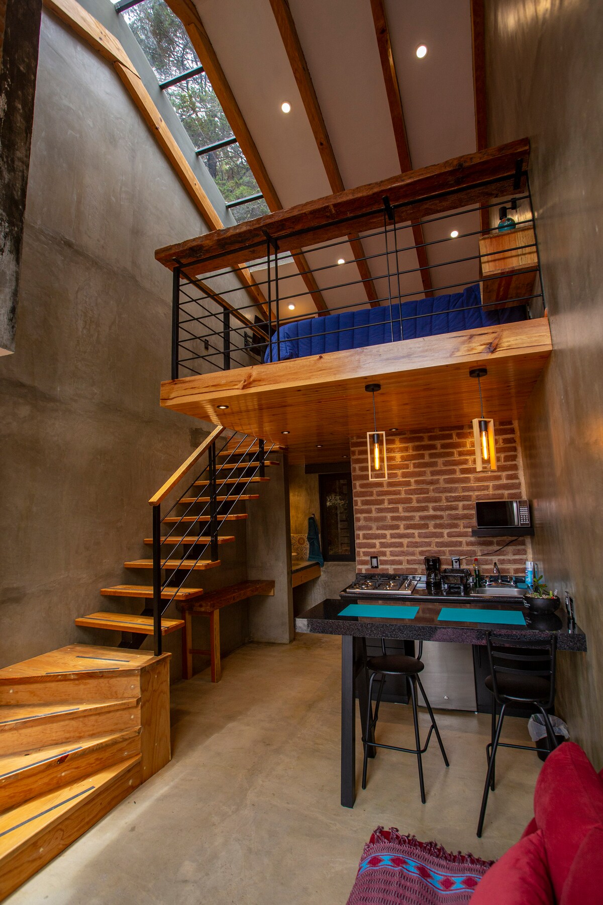 A modern loft space features a double-height ceiling with large windows allowing natural light to filter in. A wooden staircase leads to a sleeping area above, while a compact kitchen and dining counter are positioned below, accompanied by black bar stools.