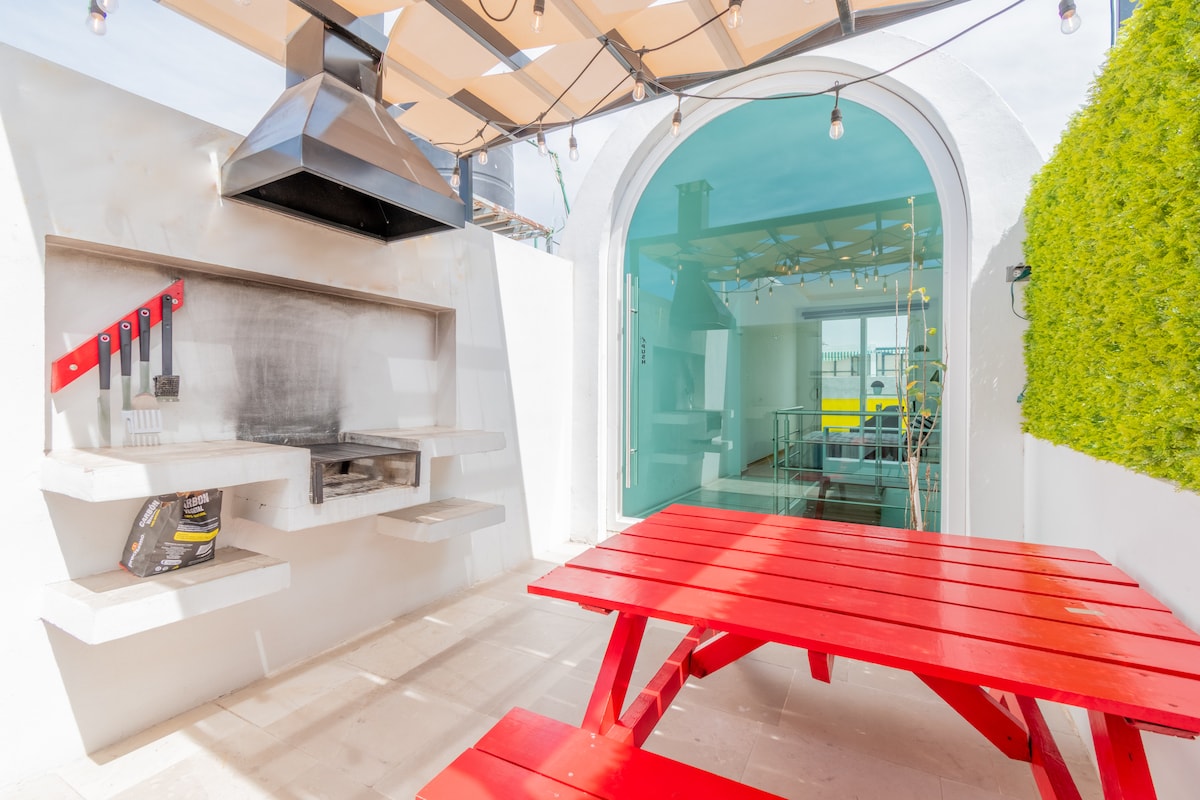 An outdoor cooking area features a built-in grill with cooking utensils neatly arranged. A vibrant red picnic table offers a space for dining. Surrounding greenery adds a touch of nature, while overhead lighting creates a warm ambiance for evening gatherings.