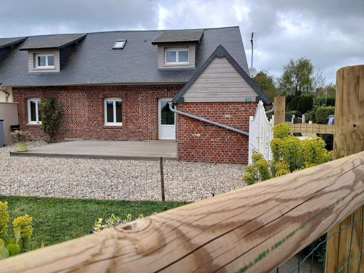 A charming brick and wood structure is depicted, featuring a sloping roof and multiple windows. A wooden fence runs along the foreground, with a gravel yard and garden plants visible. The entrance is marked by a small staircase leading to the door.